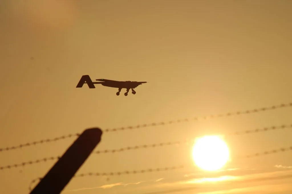 Silhouette of a Windracers autonomous cargo drone flying across a sunset sky, with barbed wire fence in the foreground.