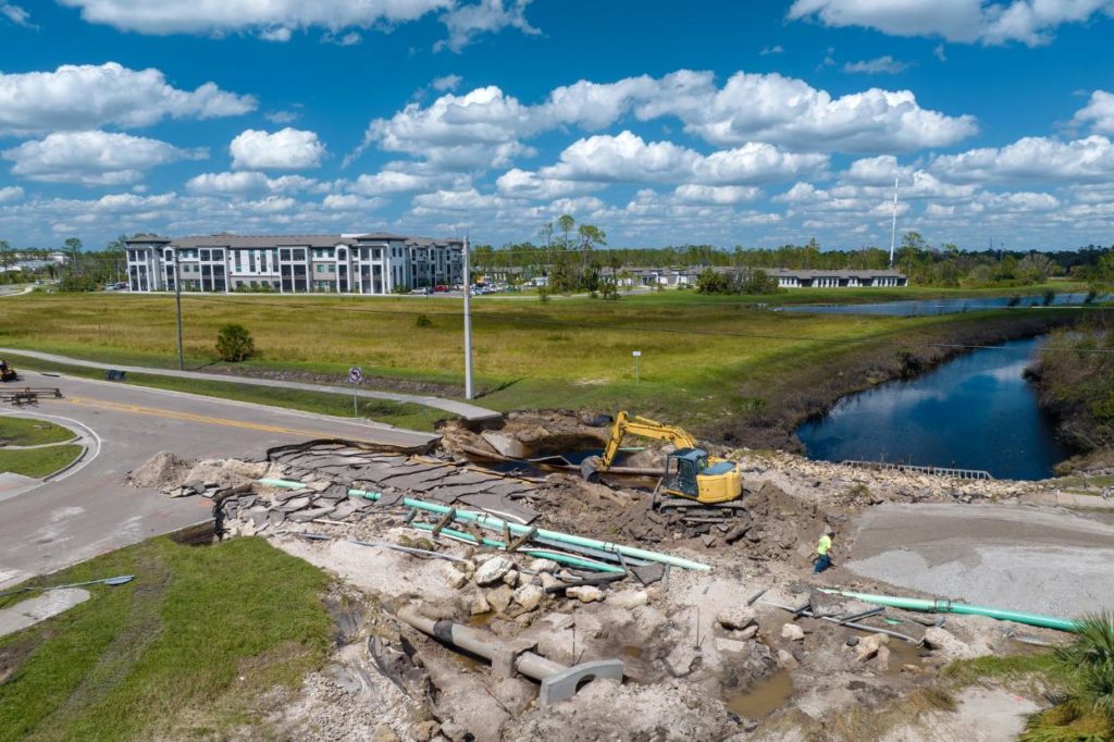 Heavy-lift logistics drones An aerial view of a severely damaged road junction, with cracked and uplifted tarmac exposing underground pipes, a yellow excavator working in the centre of the damage, and a residential building, waterway and open grassland visible in the background under a blue sky.