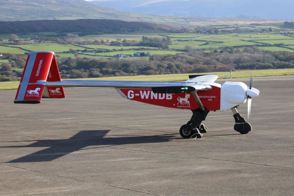integrate autonomous drones for disaster logistics A red and white Windracers Ultra uncrewed aircraft, registration G-WNDB, parked on an airfield runway with rolling green hills and farmland visible in the background.